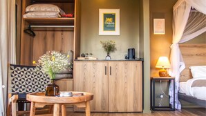 A hotel room with a bed, a wooden wardrobe, a desk with a chair, a small table with a vase of flowers, and a nightstand with a lamp. - Studio Dwelling (Tigoni)