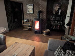 A cozy room with a wood-burning stove, a brick wall, a shelf with bottles, and a couch. - Lovely summer house in Marielyst (Væggerløse)