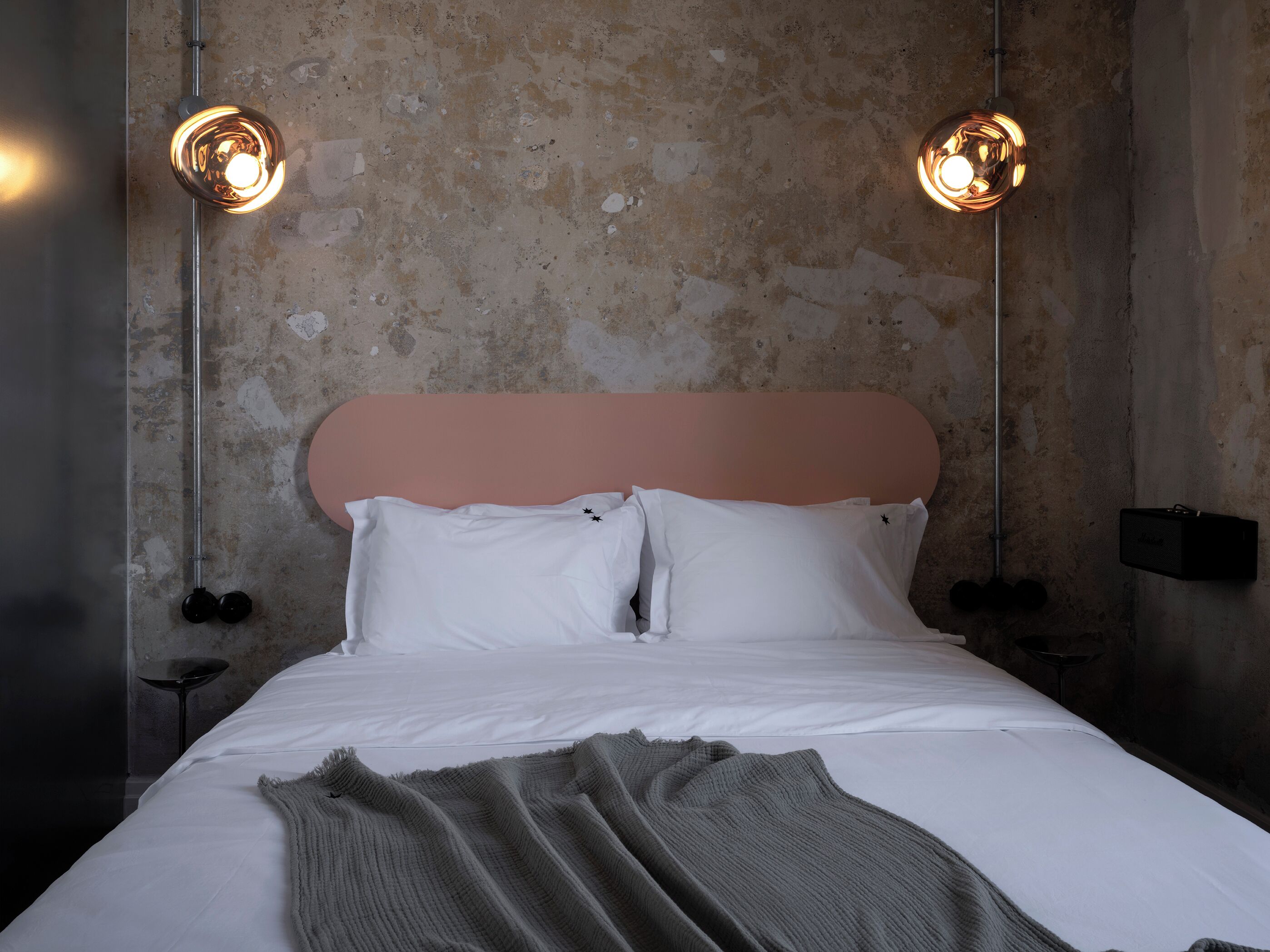 A bed with white linens, a grey throw blanket, and a pink headboard. Two wall-mounted lights are visible.
