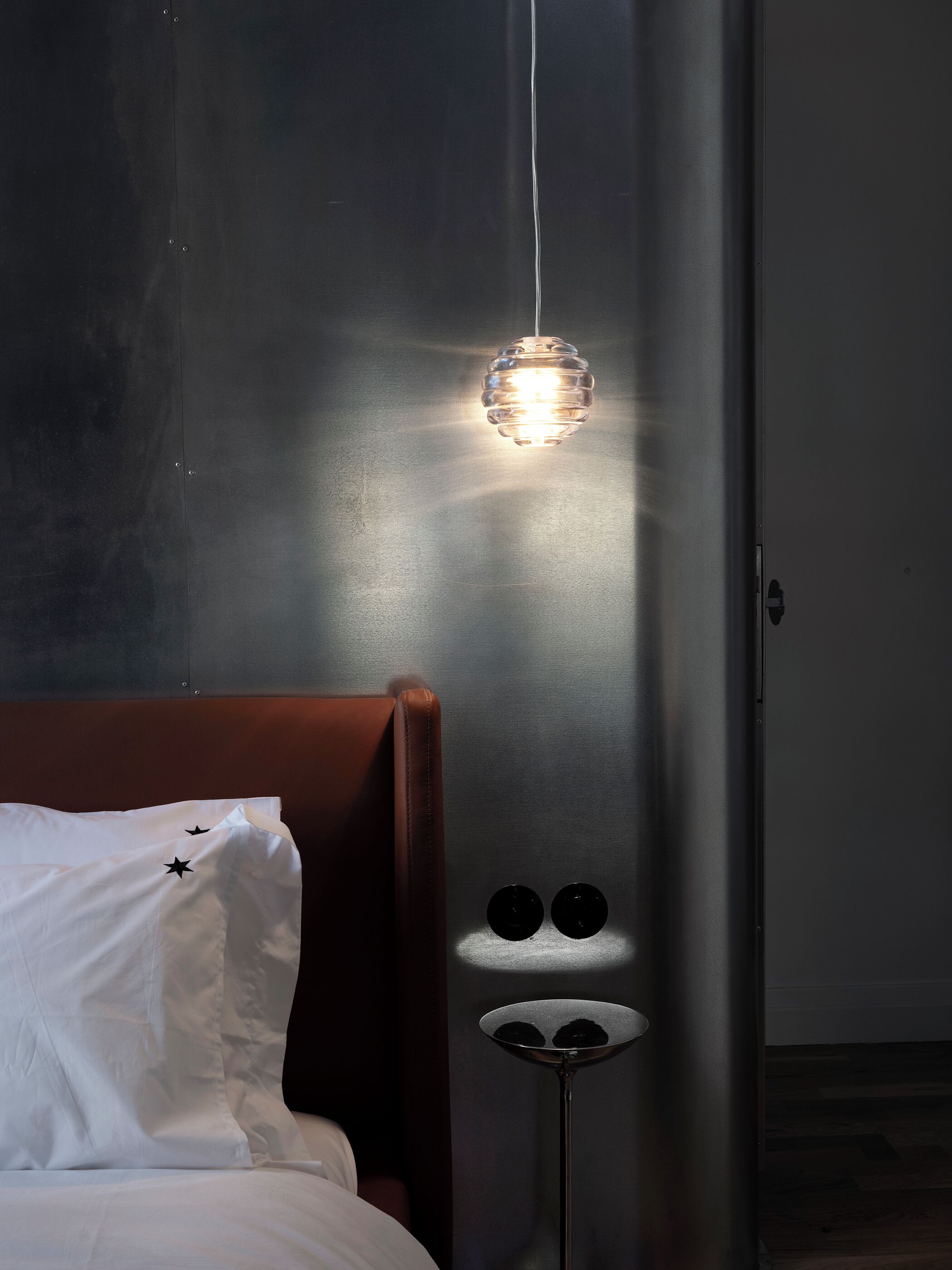 Een slaapkamer met een bed, een nachtkastje met twee zwarte voorwerpen, een hanglamp en een deur.