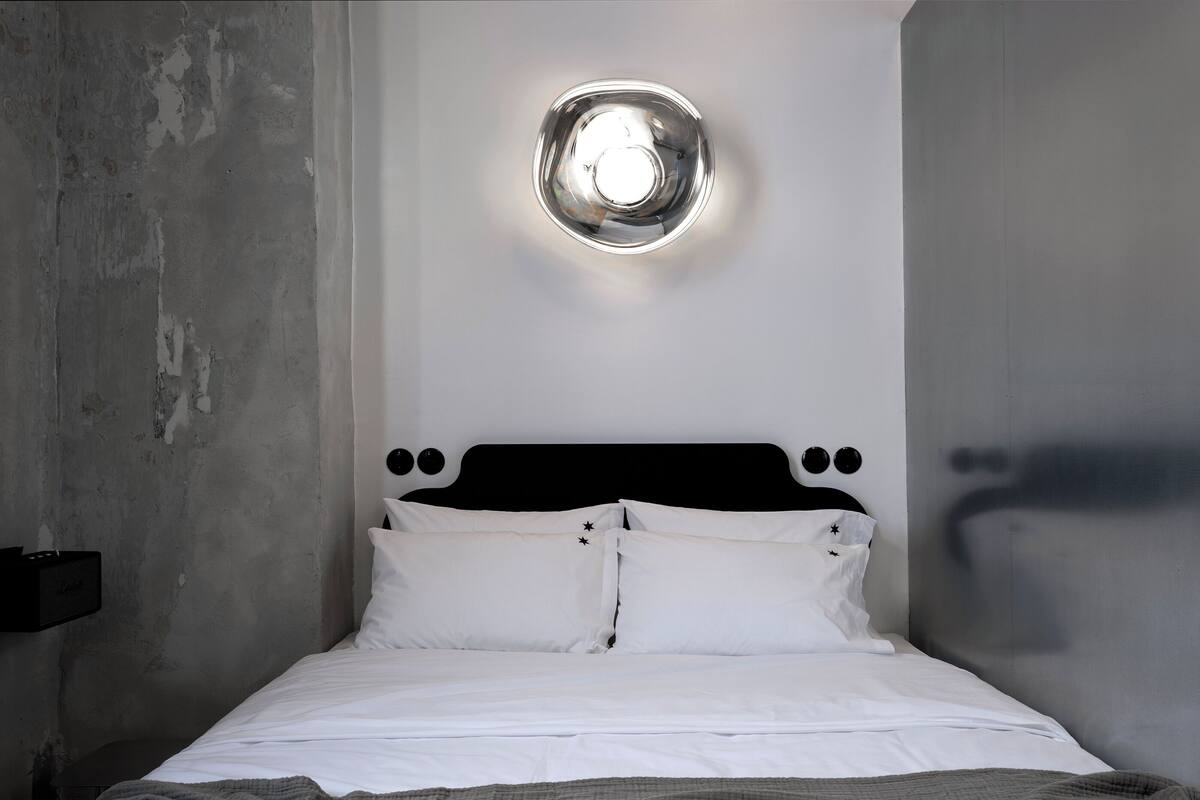 A bed with white bedding and a gray blanket, a black headboard, and a round light fixture on the wall.
