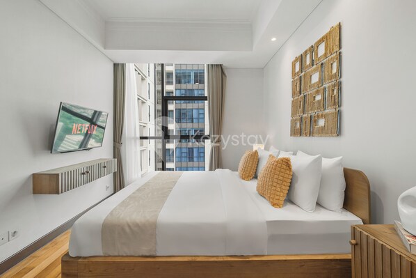 A modern hotel room with a large bed, a mounted TV, a view of the city, and a wooden headboard.