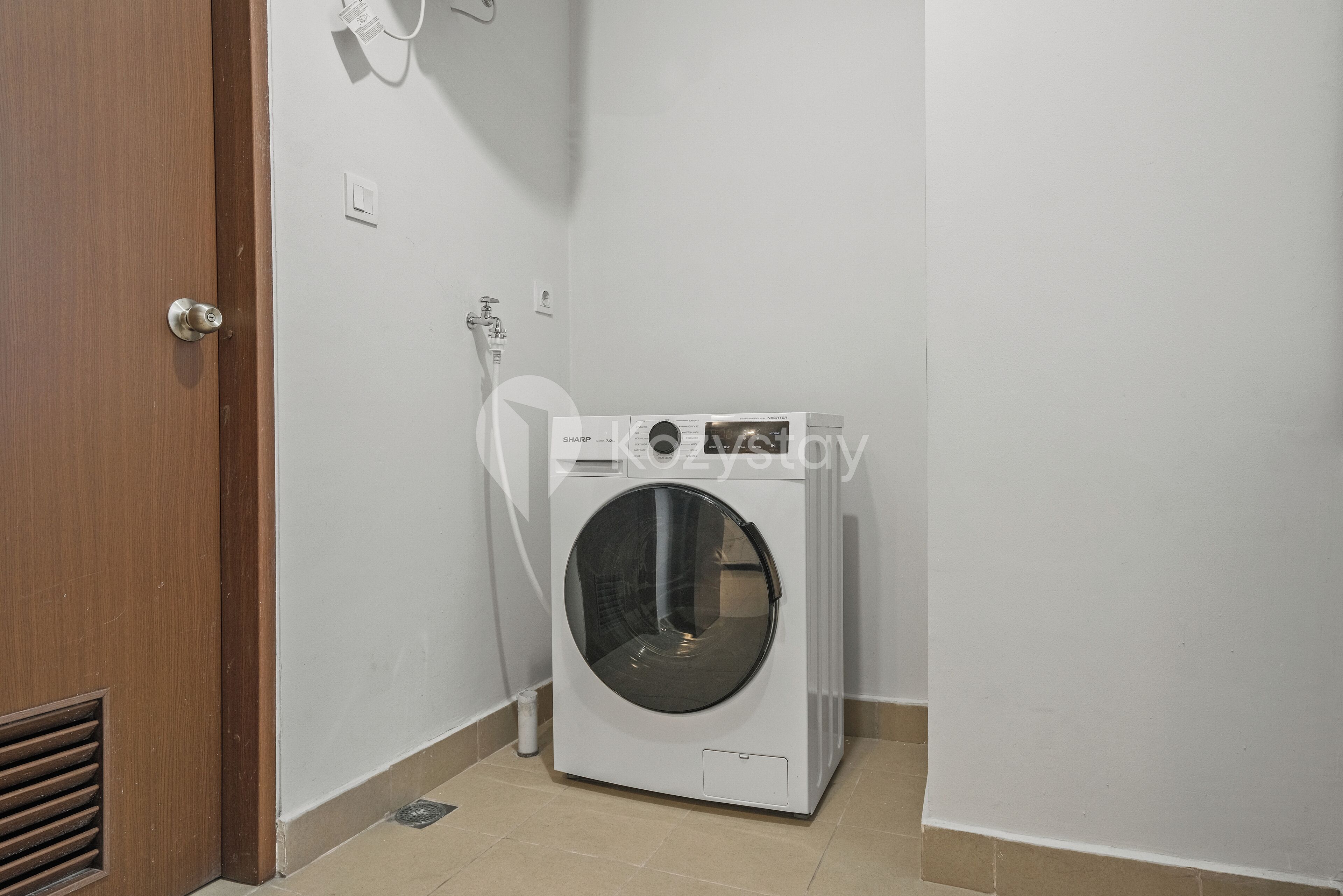 A compact, white washing machine in a small laundry room with a wooden door and a white wall.