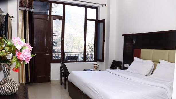 A bedroom with a large bed, a chair, a small table with a cup, and a window with curtains. - Hotel Hillora Mcleodganj (Dharamshala)