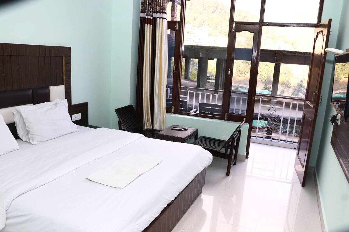 A hotel room with a large bed, a small table, a chair, and a balcony with a view of the outdoors.