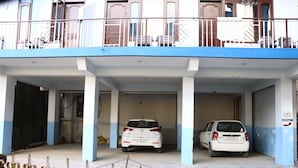 A parking area with two white cars parked under a blue and white building. - Hotel Hillora Mcleodganj (Dharamshala)