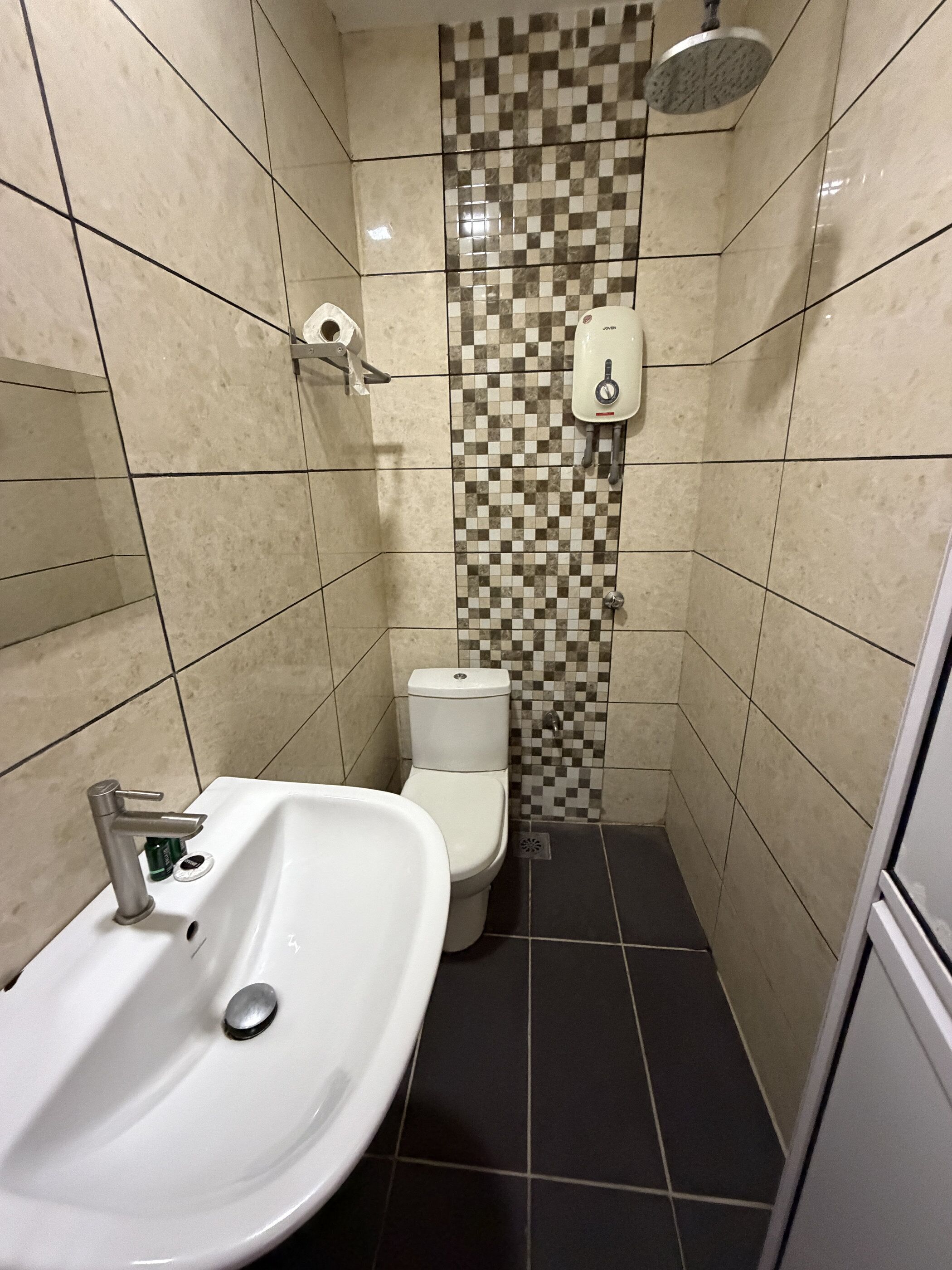 Une petite salle de bain avec un lavabo, des toilettes et une douche, le tout de couleur blanche. Les murs sont recouverts de carreaux disposés en motif à carreaux.