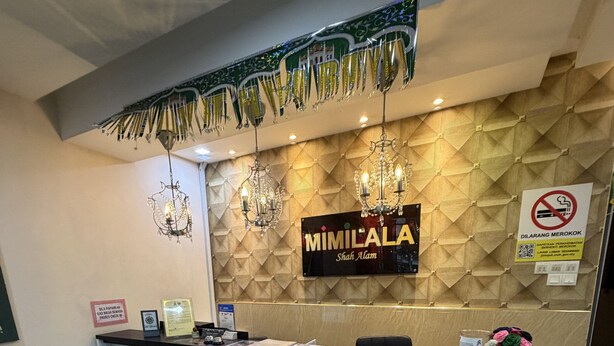 Een receptiebalie met een bord waarop 'MIMILALA Shah Alam' staat, een kroonluchter, een rookvrij bord en een bank met een rode kussen.