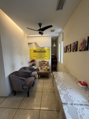 Ein Hotelzimmer mit Sofa, Sessel und einem Bett mit gemustertem Bettbezug. Ein Deckenventilator ist sichtbar, und ein Schild mit der Aufschrift 'Mimilala Hotel & Homestay' befindet sich an der Wand.