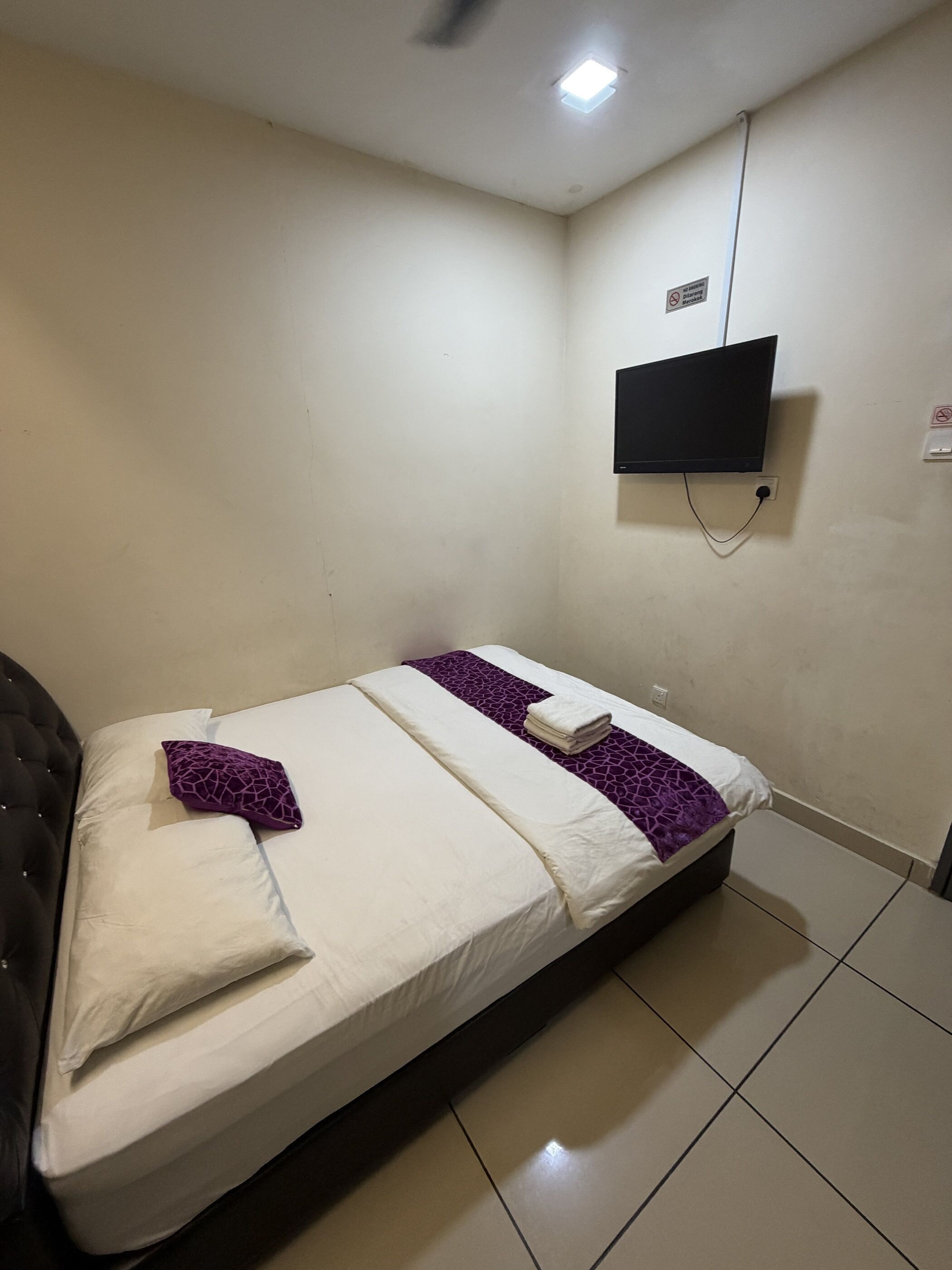 Una cama individual con colcha blanca y morada, cabecero negro, televisor de pantalla plana en la pared y un enchufe.