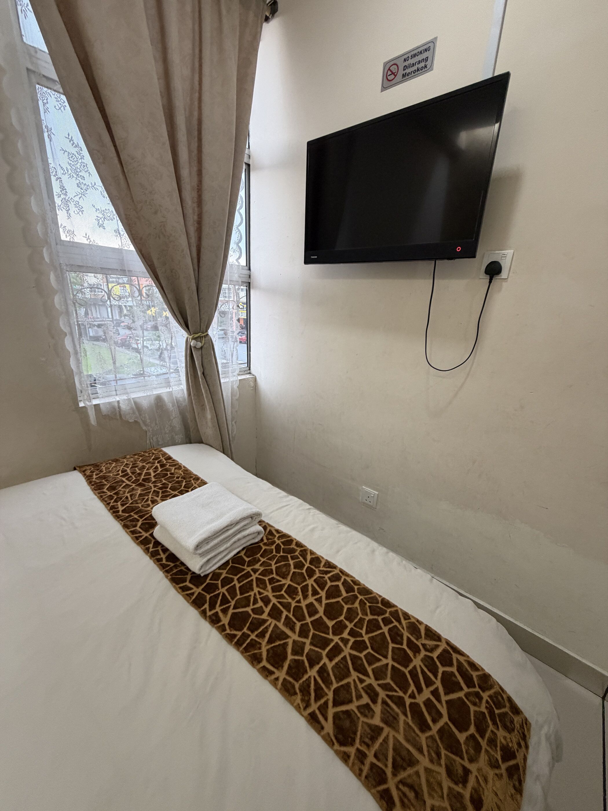 Quarto de hotel com cama, televisão fixada na parede, janela com cortinas e placa indicando proibido fumar.
