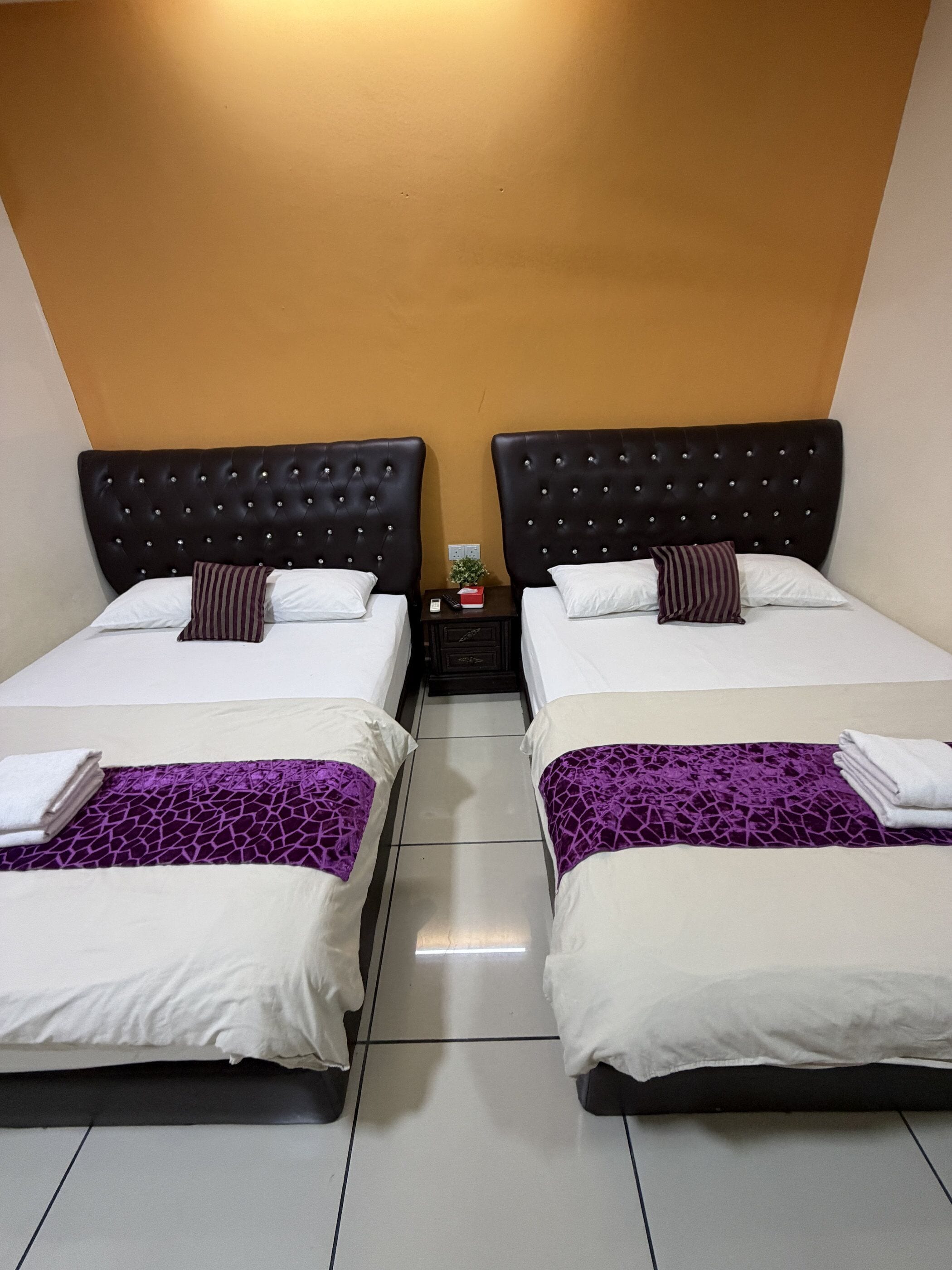 Habitación de hotel con dos camas, cada una con cabecero, una mesita de noche pequeña y una colcha morada estampada.