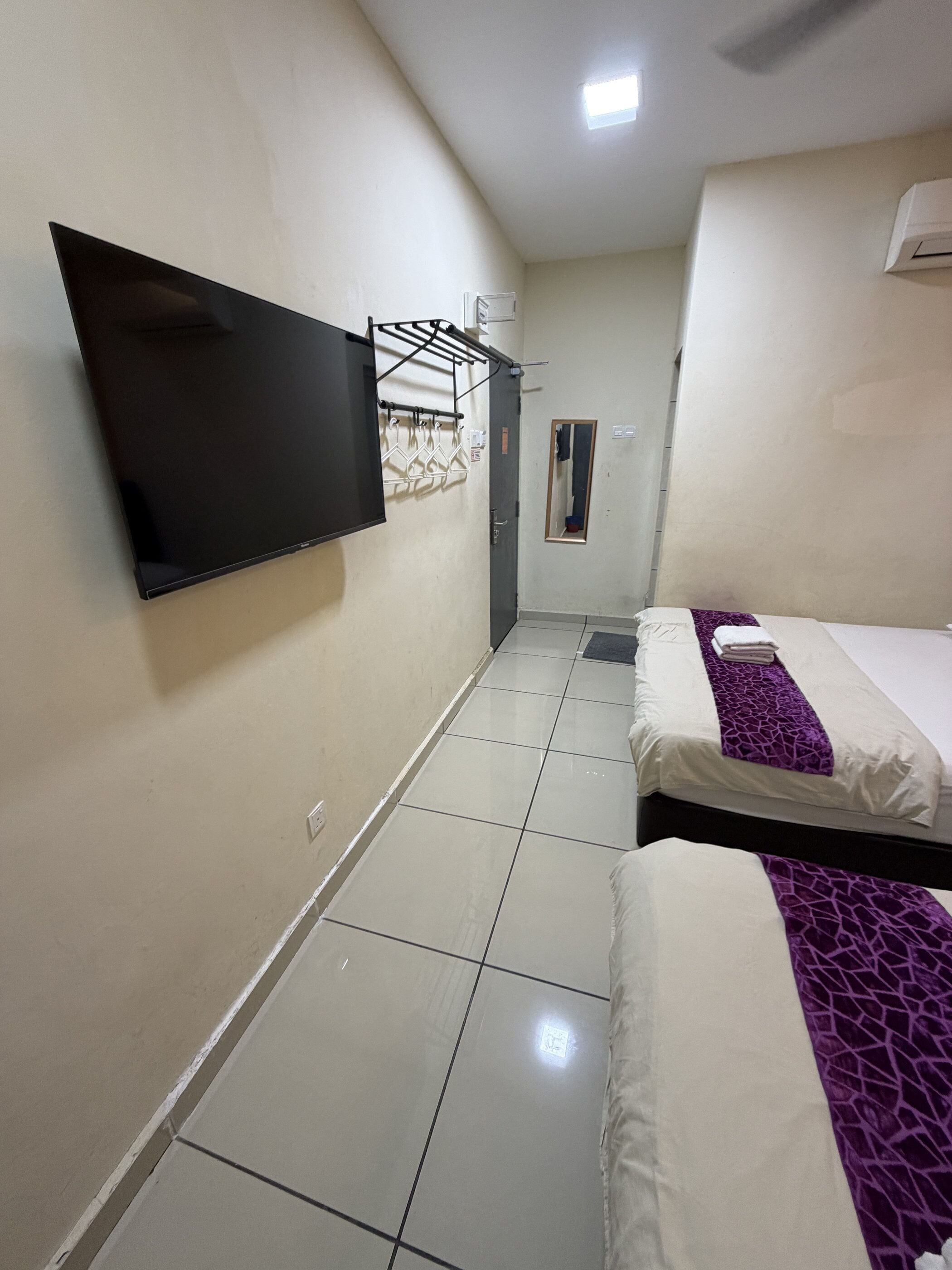 A hotel room with a flat-screen TV mounted on the wall, a bed with a purple patterned bedspread, and a tiled floor.