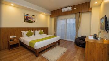 A hotel room with a wooden bed, a desk, a chair, a nightstand, a window with curtains, and an air conditioning unit.