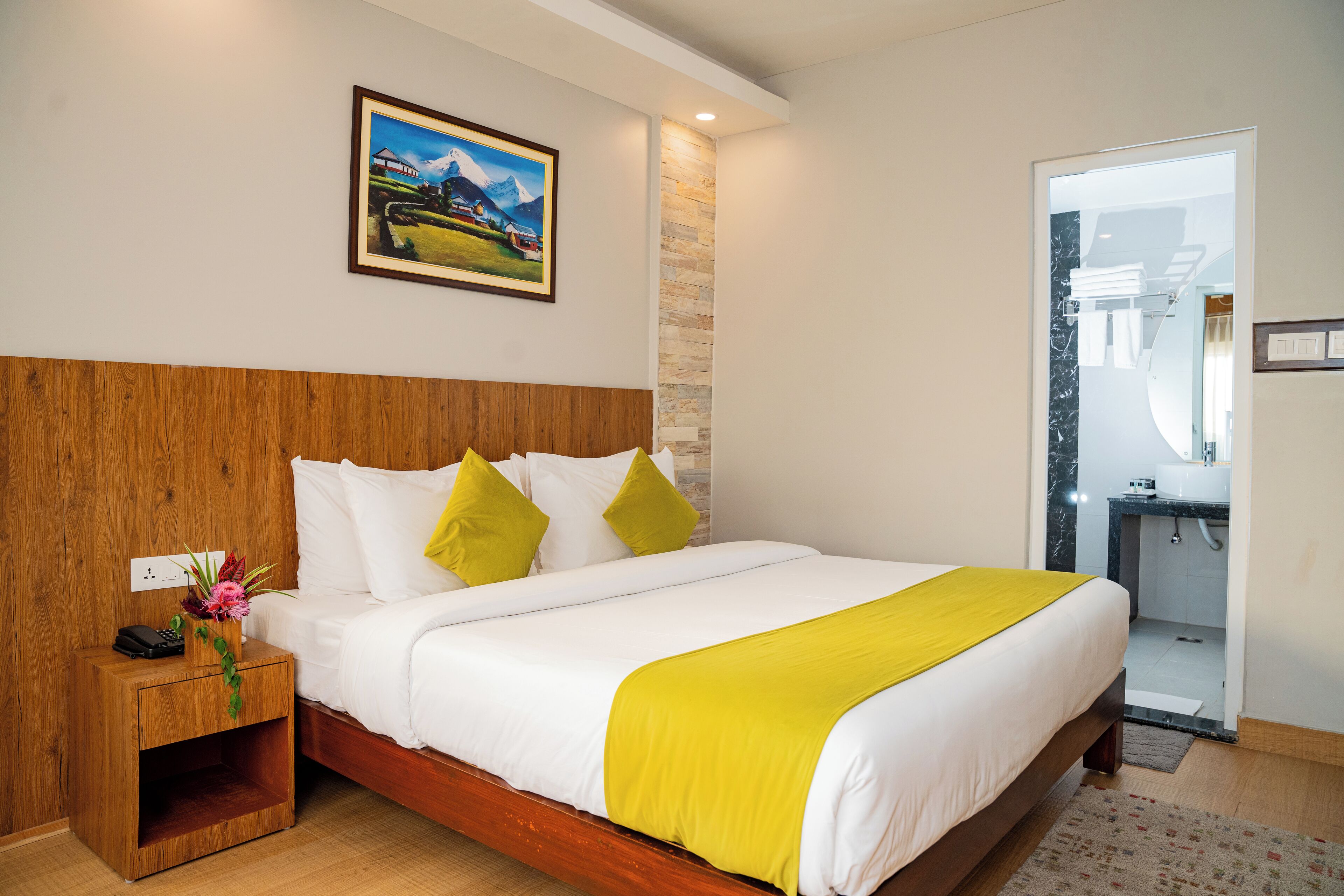 Una habitación de hotel con cabecera de madera, una cama con ropa blanca y un plaid amarillo, una mesita de noche con un teléfono y un florero, y un cuadro de un paisaje montañoso en la pared.