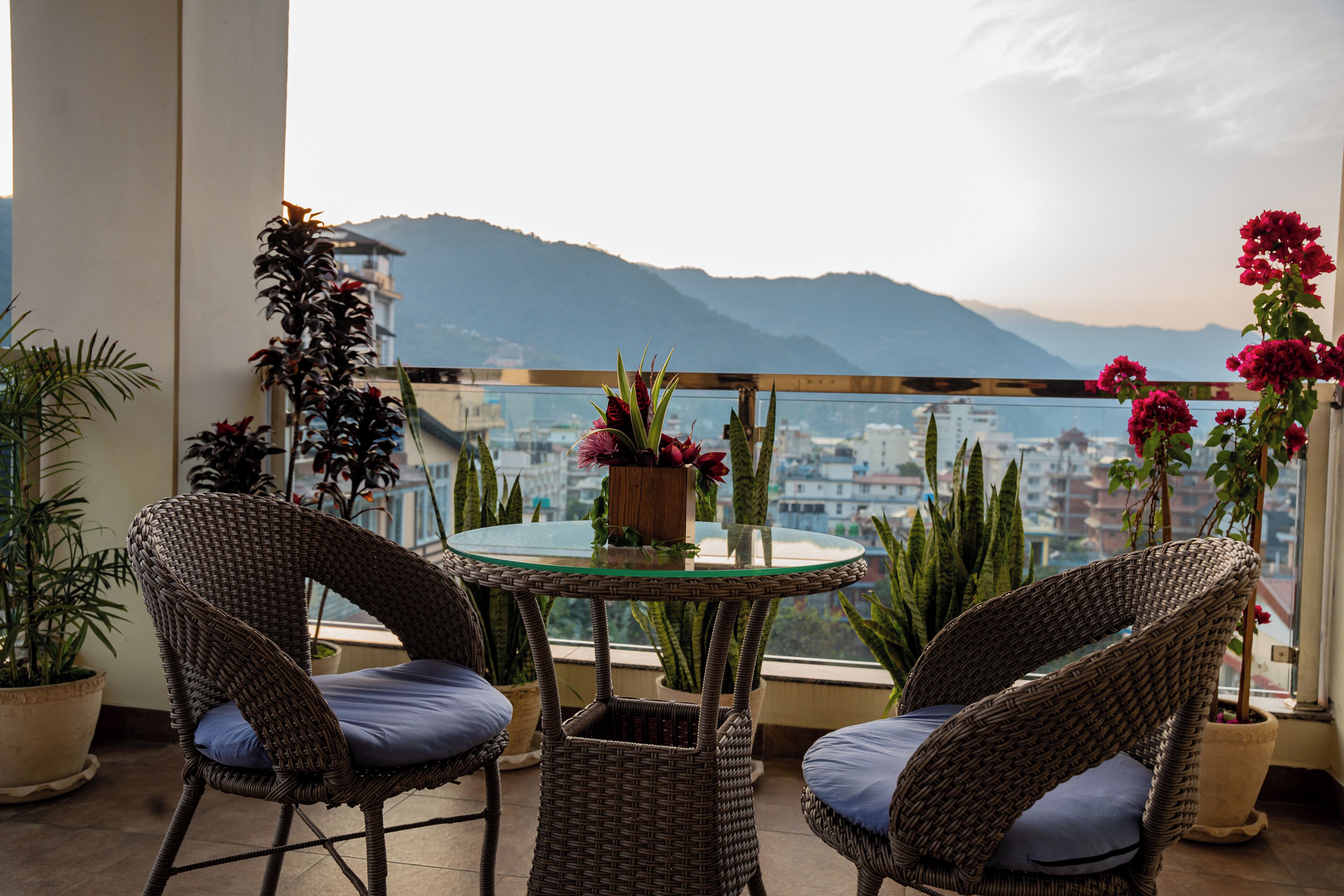 A balcony with wicker chairs and a glass table, overlooking a cityscape and mountains.