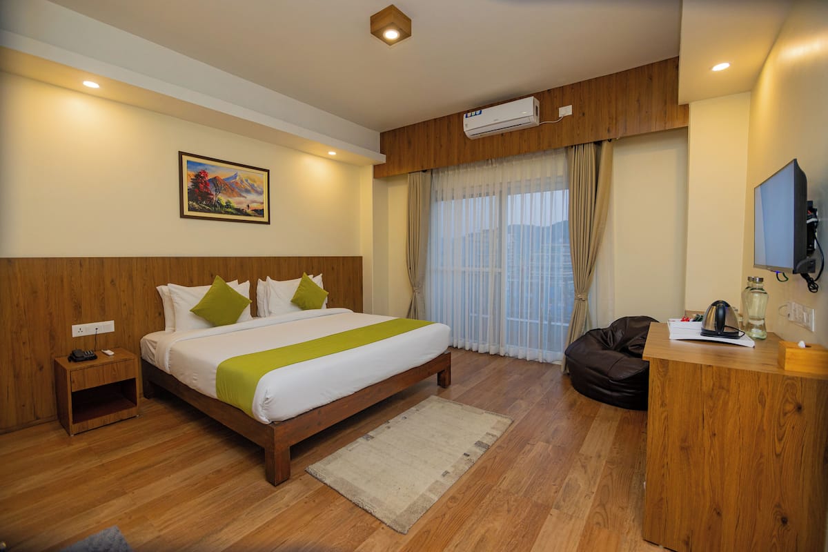 A hotel room with a wooden bed, a desk, a chair, a painting on the wall, and a window with curtains.
