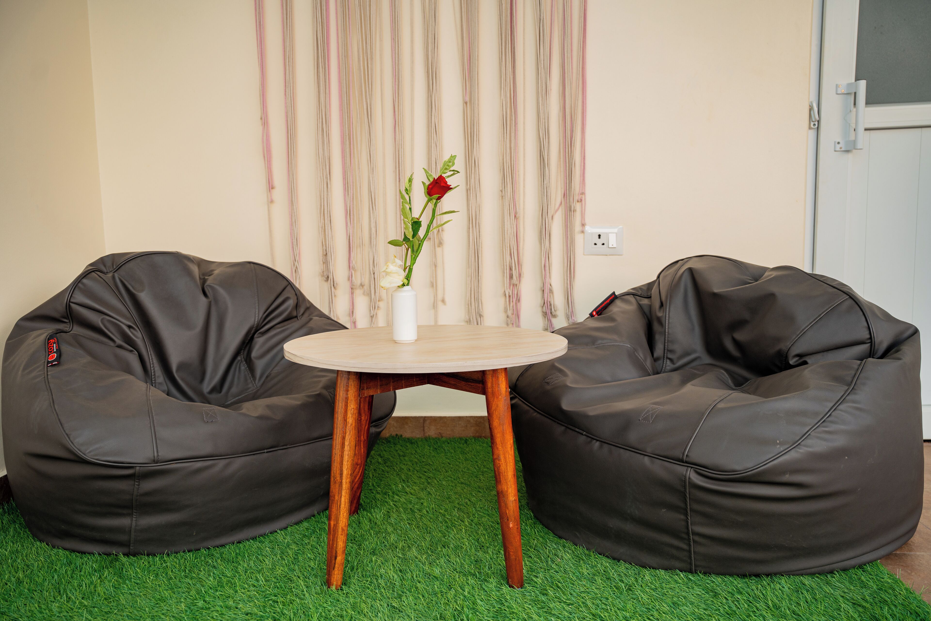 Um quarto com dois pufts pretos, uma mesa redonda pequena e um vaso com uma rosa vermelha.