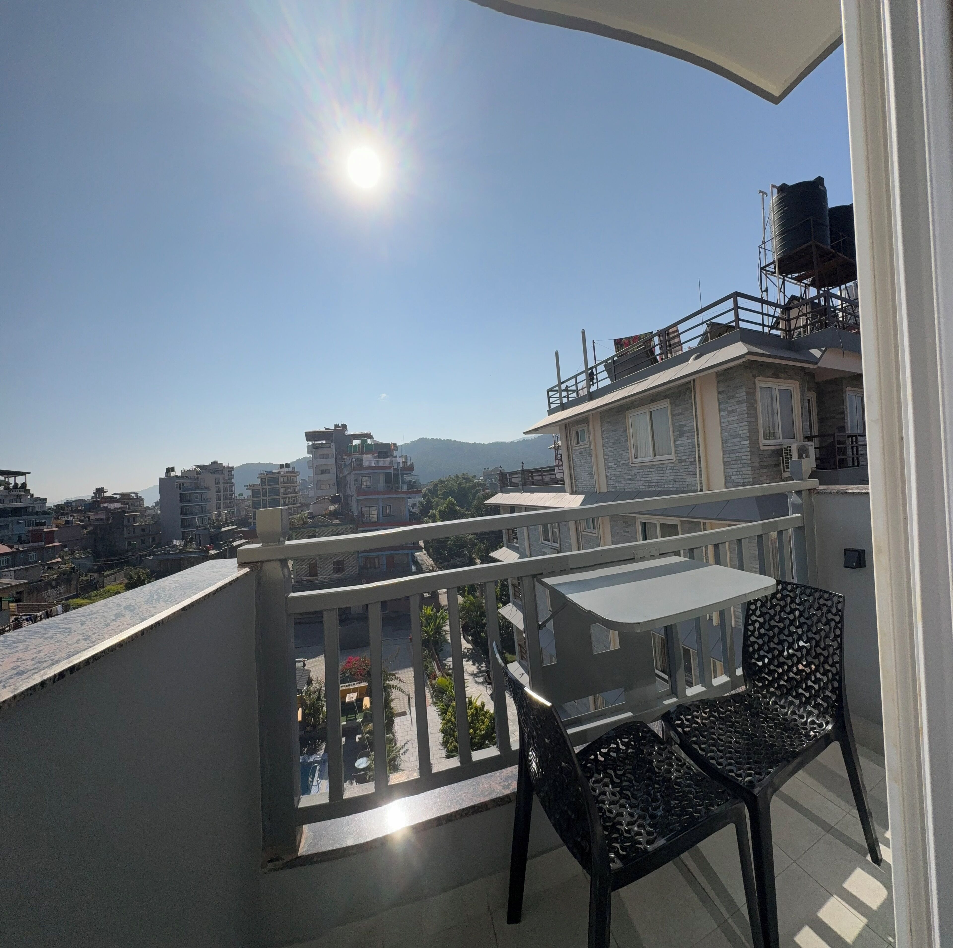 A balcony with a table and chairs, overlooking a cityscape with buildings and mountains.