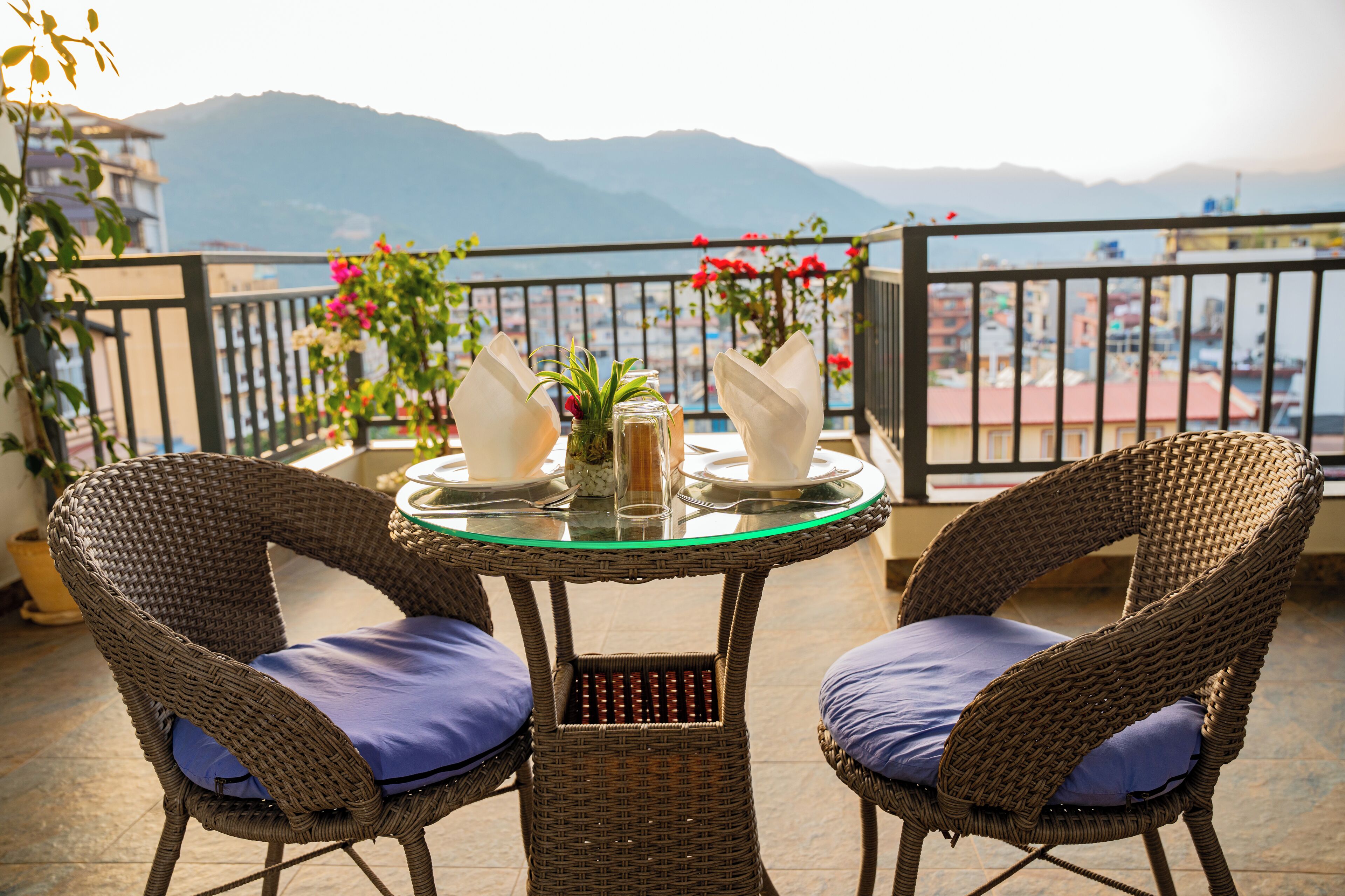 A wicker table with two chairs, a glass top, and a view of mountains and a city in the background.