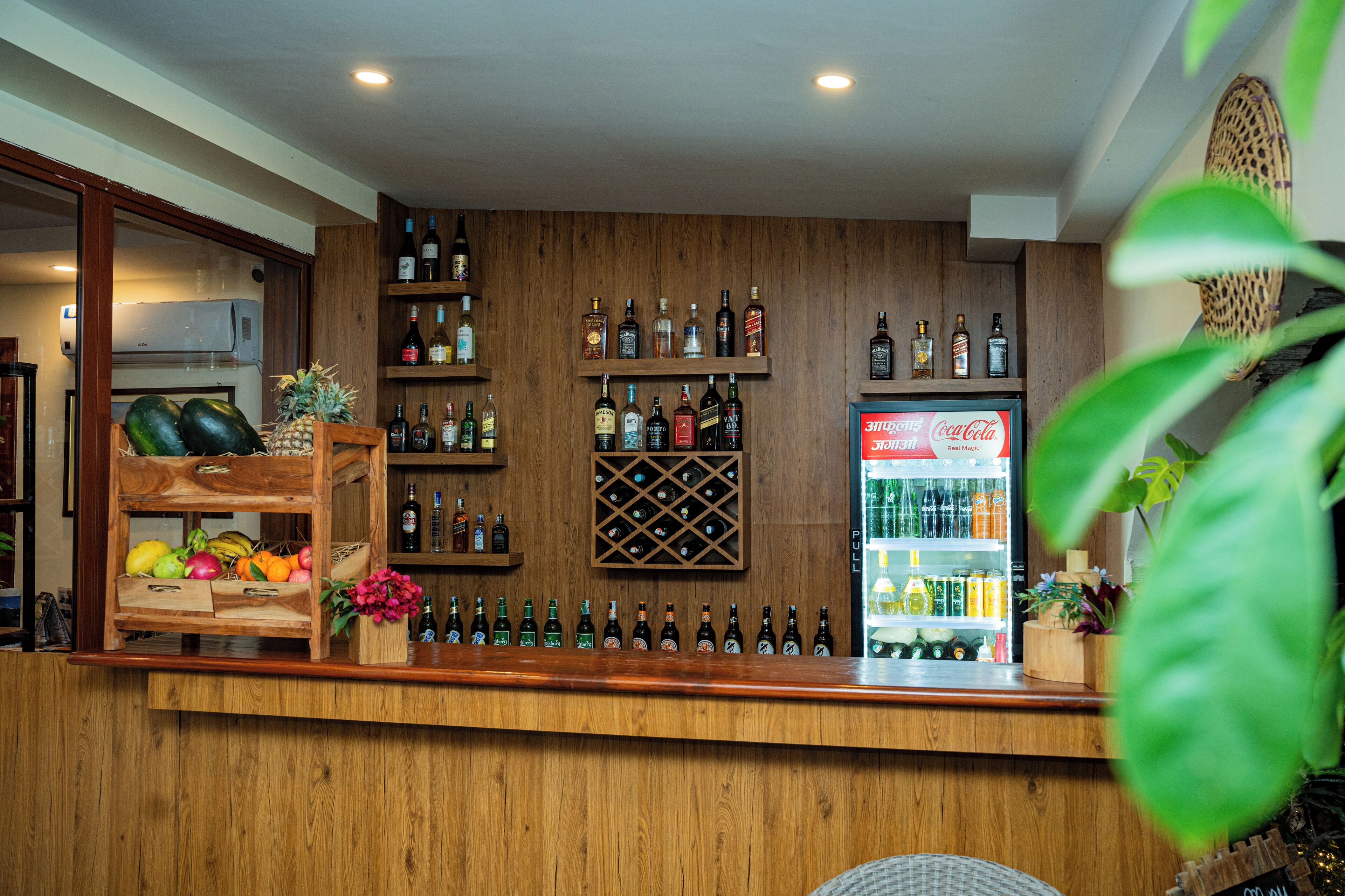Un espace bar avec des étagères en bois où sont exposées différentes bouteilles, un réfrigérateur rempli de boissons et un présentoir de fruits.