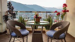 A balcony with wicker chairs and a glass table, overlooking a cityscape and mountains.