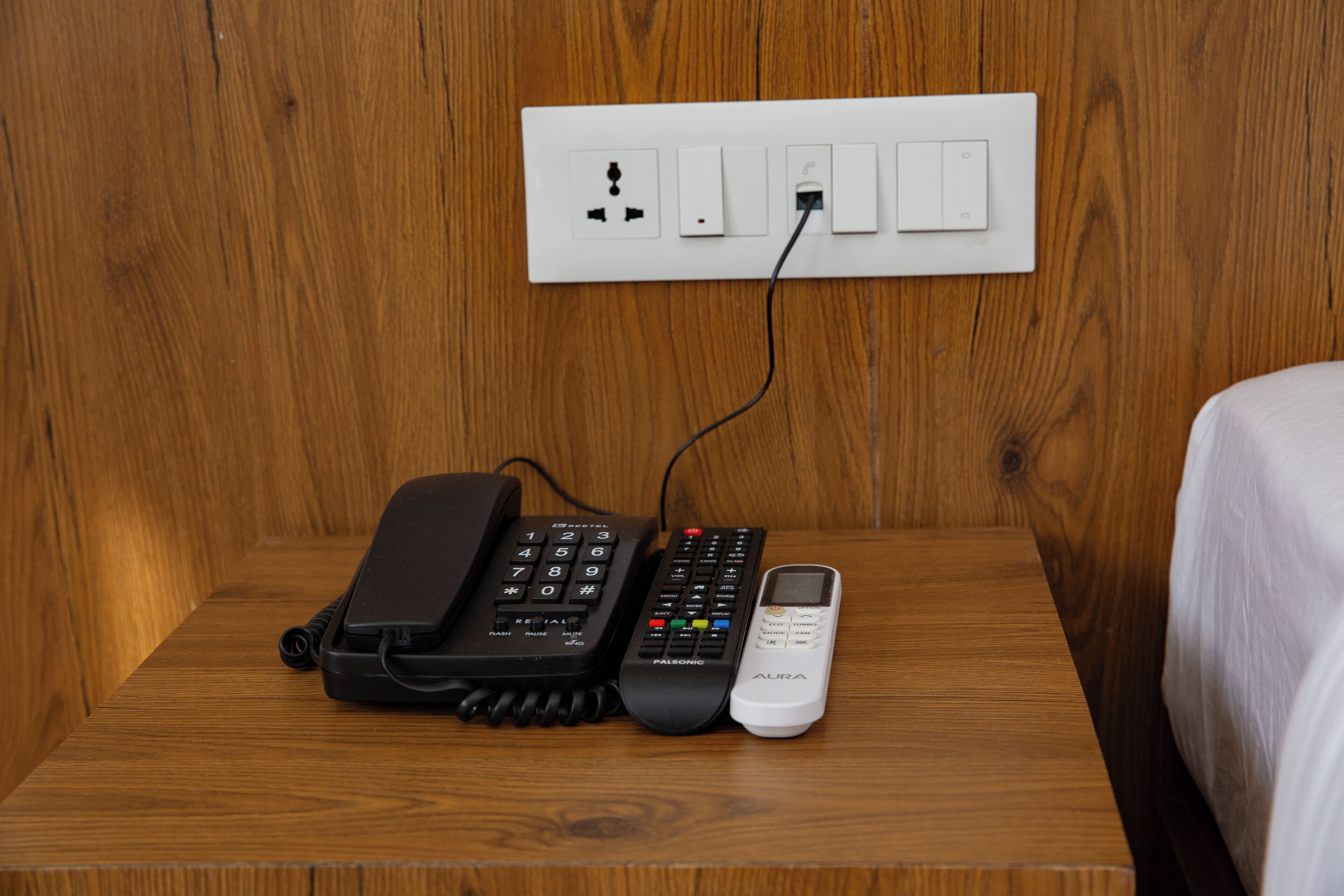 A wooden nightstand with a black telephone, a red and black remote control, and a white mobile phone.