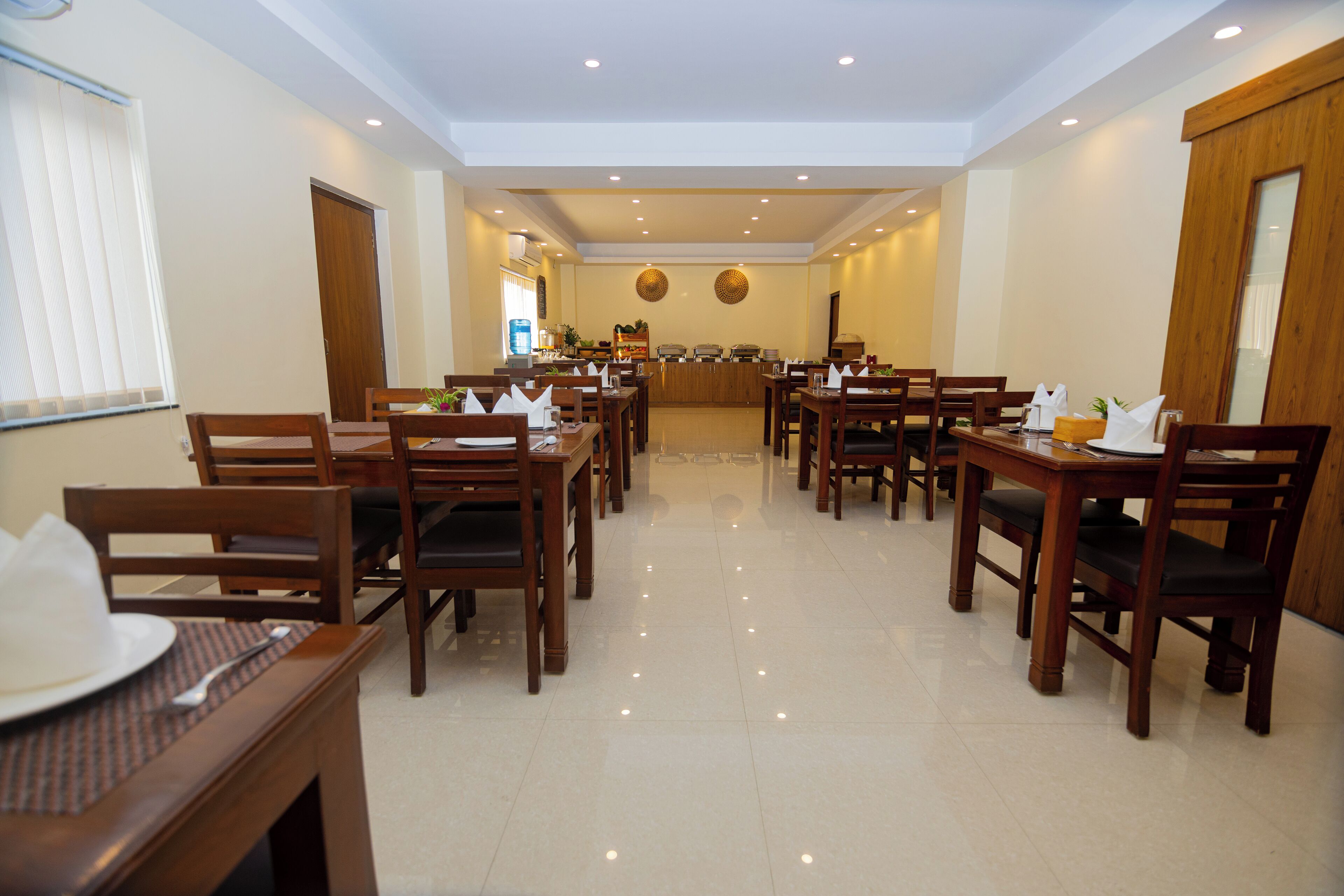 A hotel dining area with multiple wooden tables and chairs, a reception desk in the center, and a clean, tiled floor.