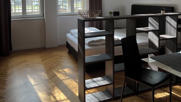 A modern room with a bed, a desk, and two windows.