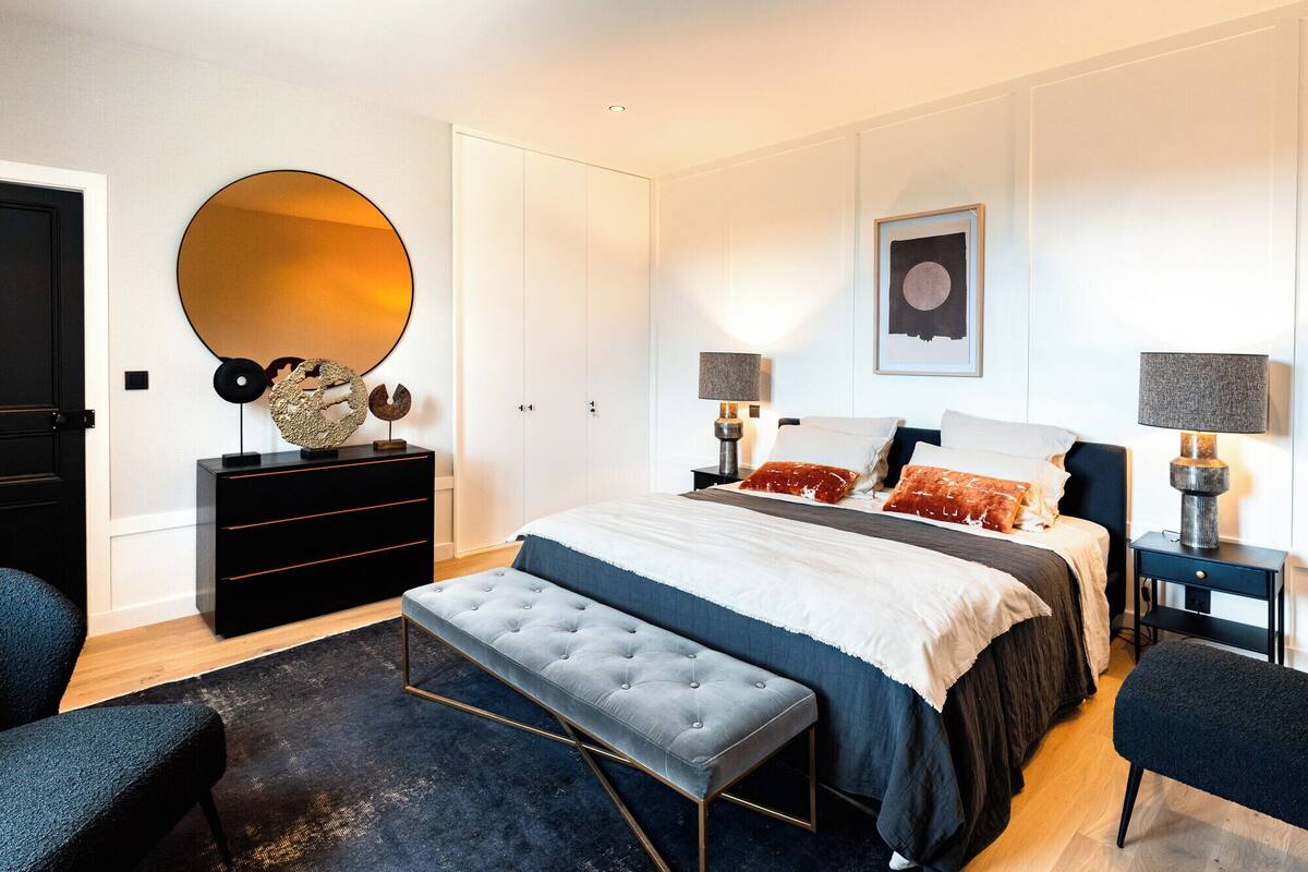 Un dormitorio moderno con una cama grande, un espejo redondo, un aparador y dos mesitas de noche con lámparas.