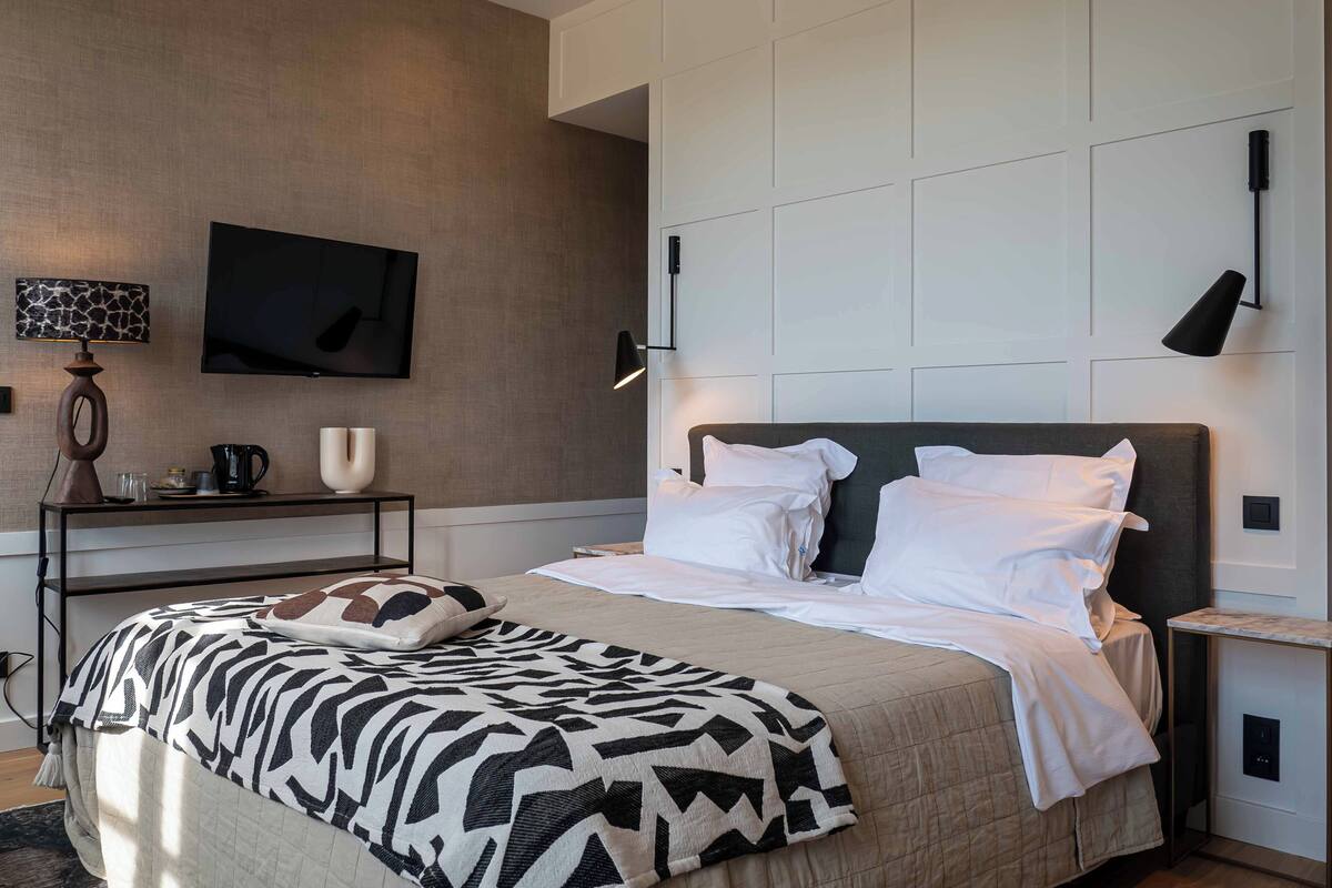 Un dormitorio moderno con una cama grande, un televisor de pantalla plana montado en la pared, lámparas de noche y una mesita con una lámpara y una cafetera.