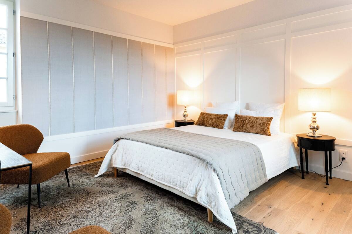 Un dormitorio con una cama grande, dos mesitas de noche con lámparas, una silla y una alfombra estampada.
