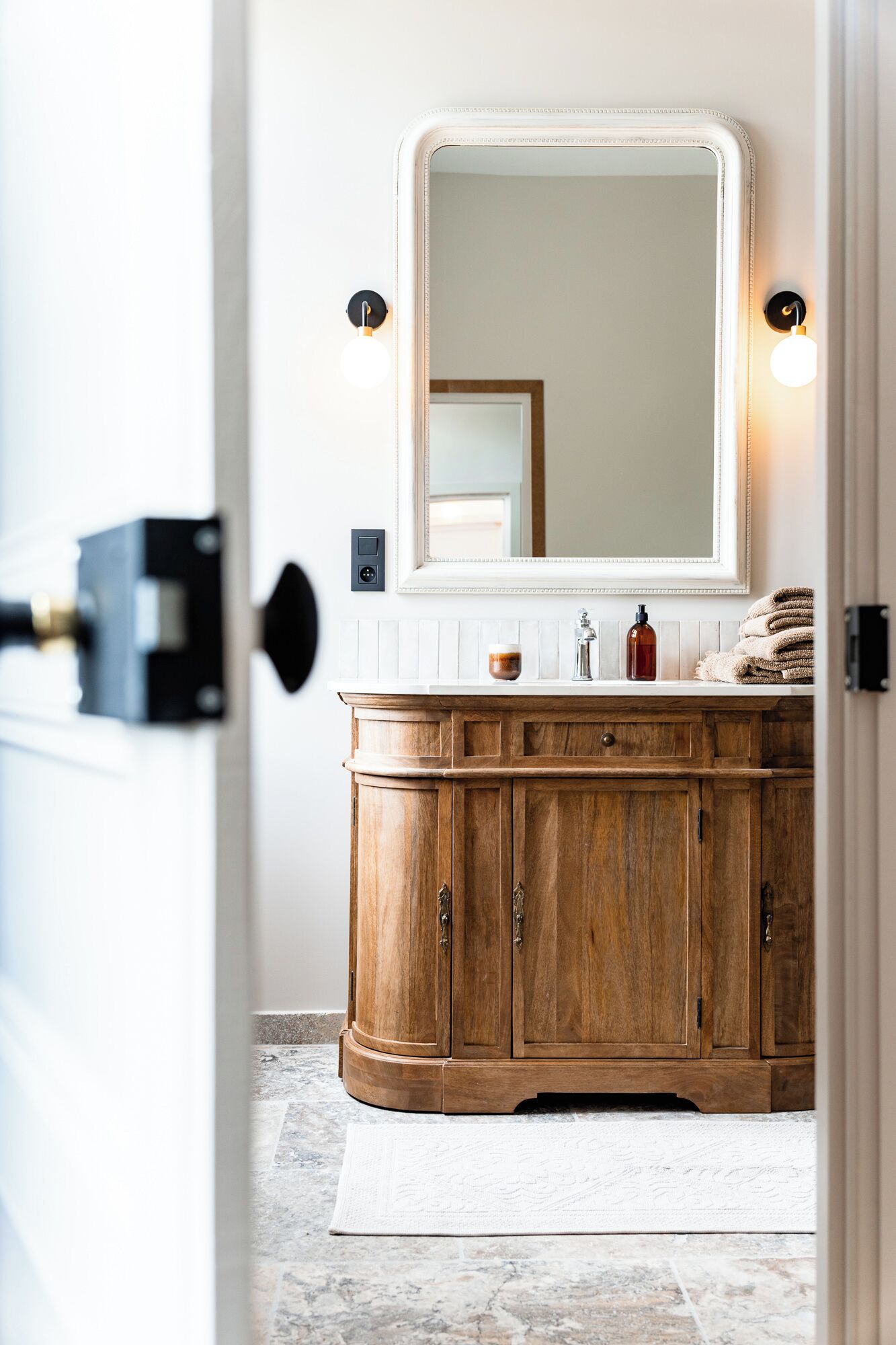A bathroom with a wooden vanity, a large mirror, and a white tiled floor.
