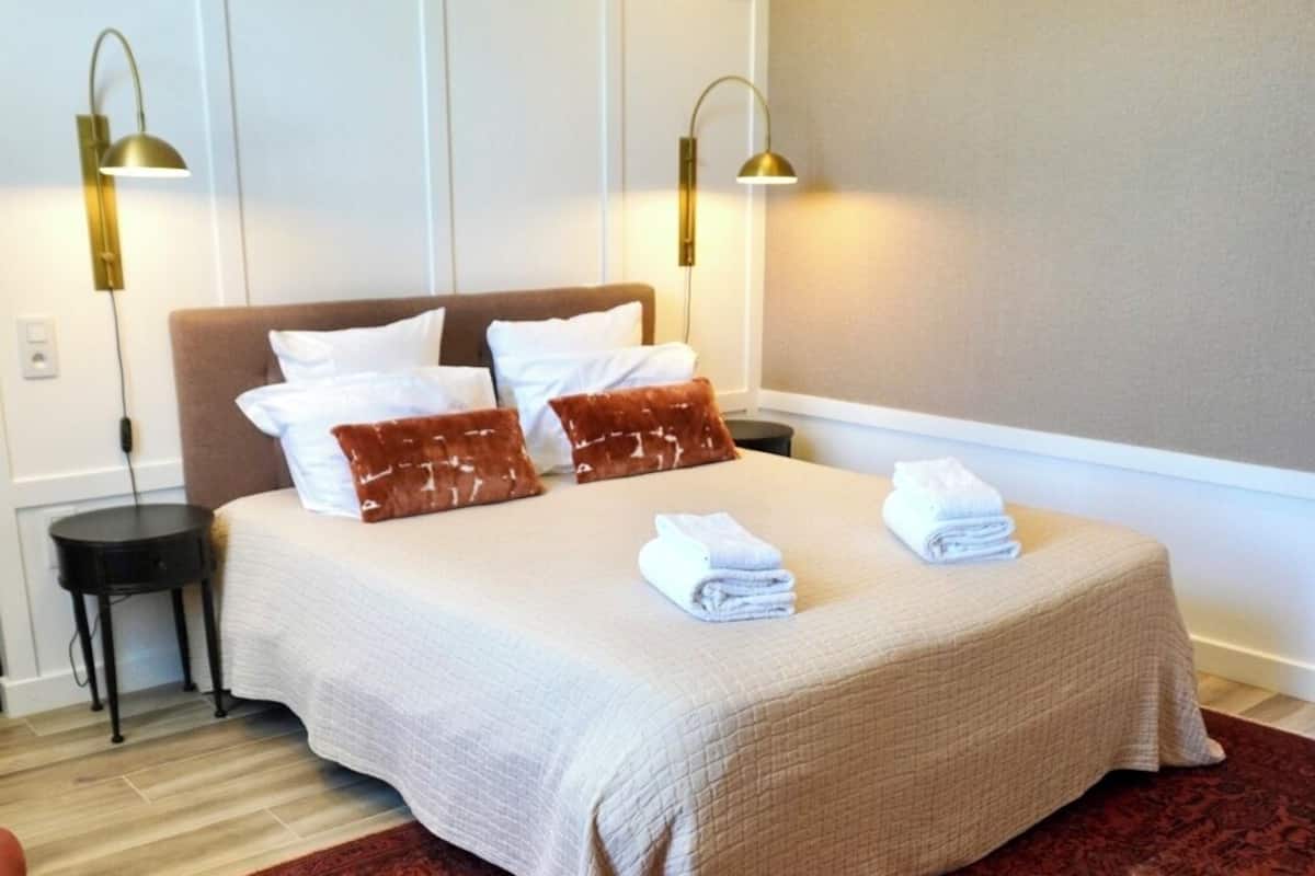 Una cama bien tendida con una colcha color beige y almohadas, dos lámparas de noche y una alfombra roja con diseño.