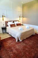 Una cama bien tendida con una colcha color beige y almohadas, dos lámparas de noche y una alfombra roja con diseño.