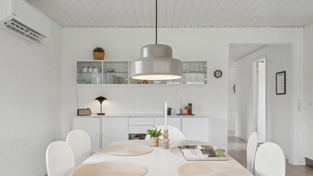 A modern dining area with a white table, four chairs, a hanging pendant light, and a white cabinet with glass doors in the background.