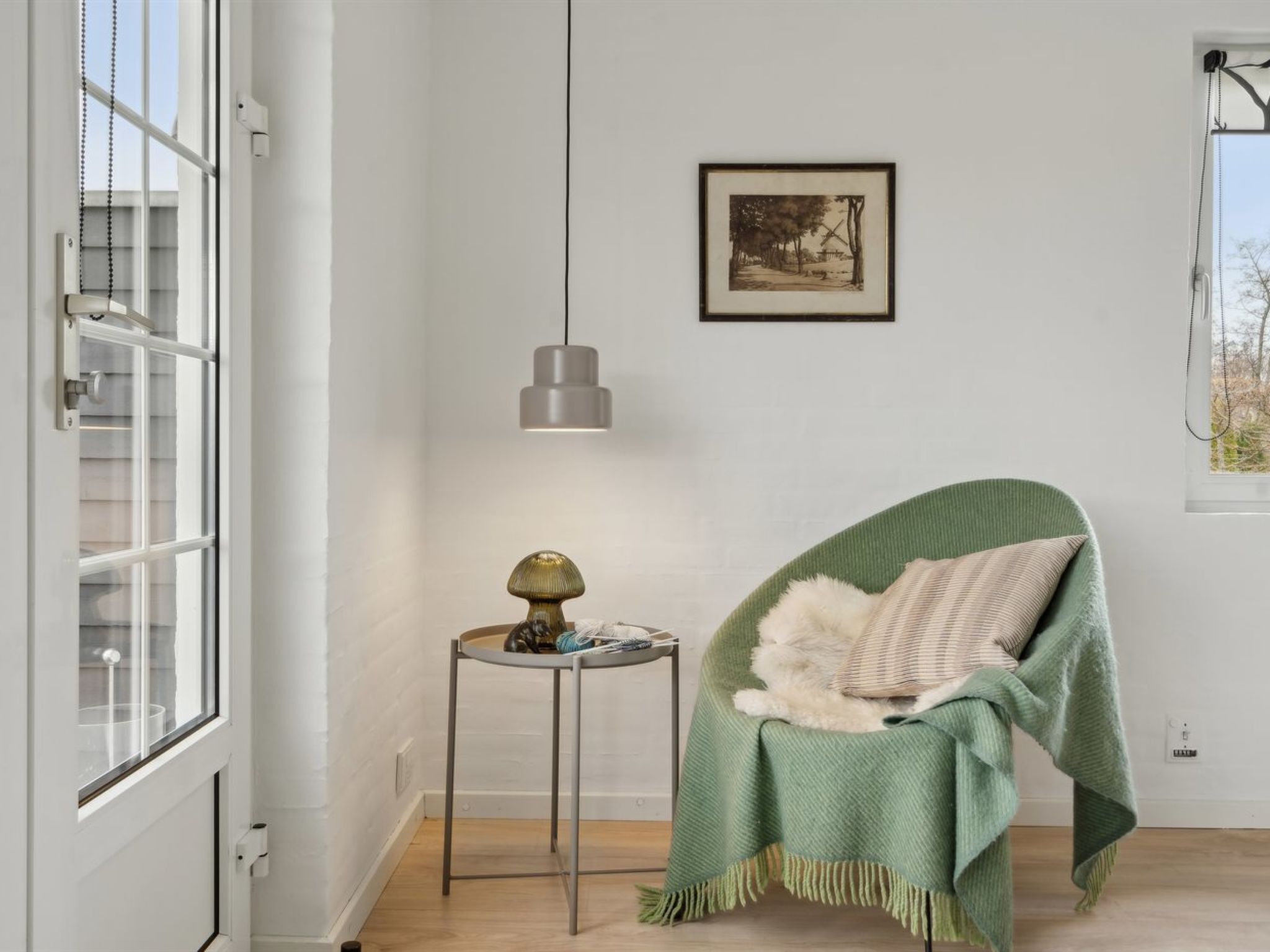 A room with a window, a framed picture on the wall, a hanging pendant light, a small table with a lamp, and a chair with a blanket.