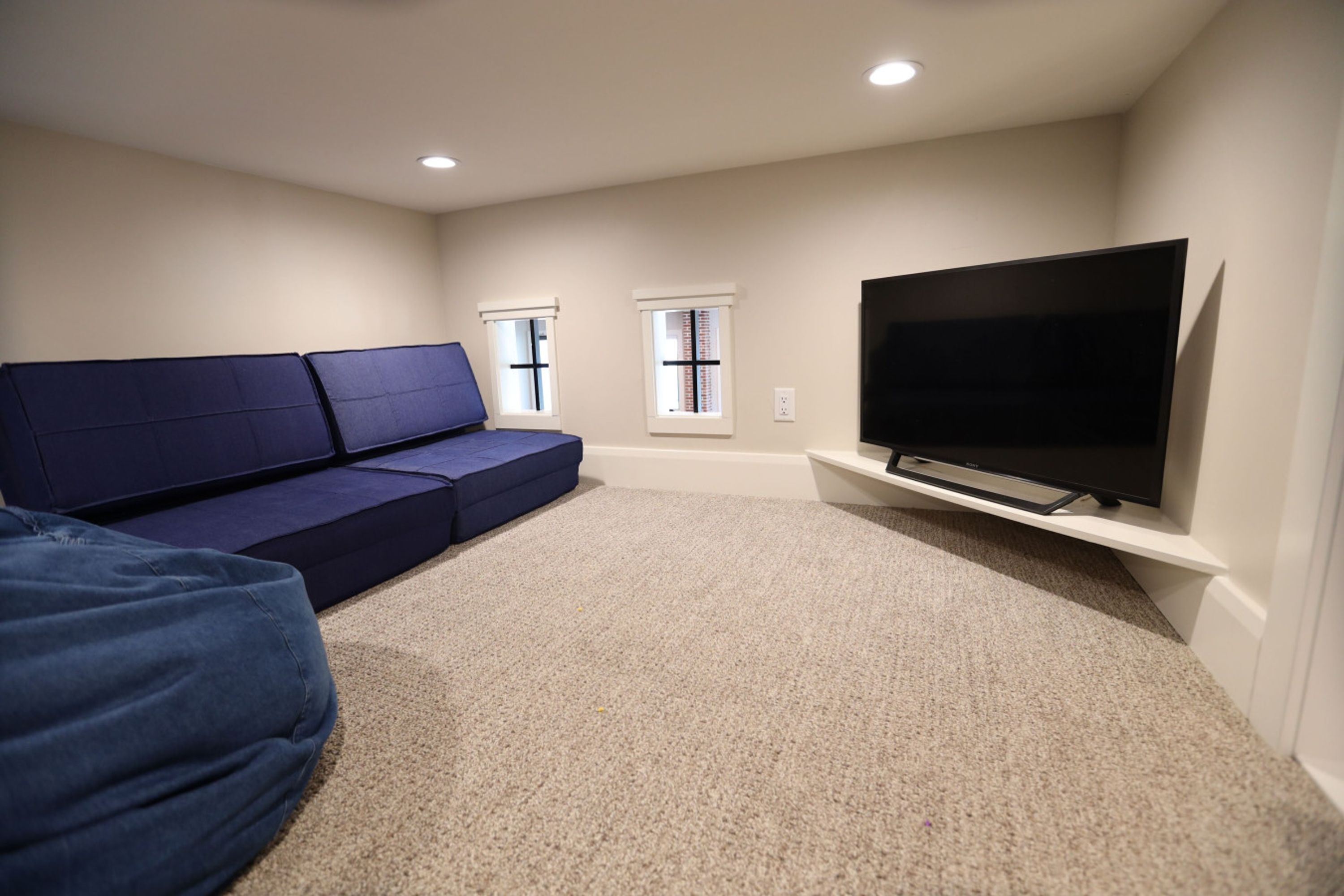 A room with a blue sofa, a flat-screen TV on a stand, two windows with blinds, and carpeted flooring.
