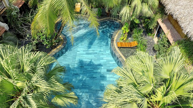 A large, clear swimming pool surrounded by lush tropical vegetation, including palm trees and other greenery. - Trang An Aroma Homestay (Ninh Binh)