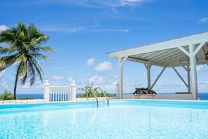A clear blue pool with a palm tree and a covered seating area. - Domaine des Tourterelles (Capesterre-de-Marie-Galante)