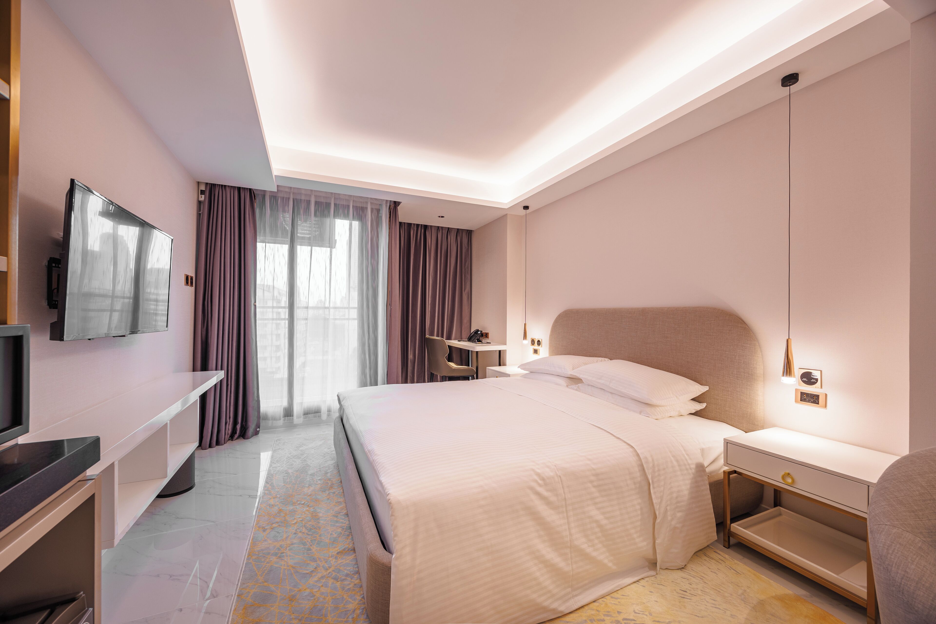 A modern hotel room with a large bed, a desk, a TV, and a desk lamp.