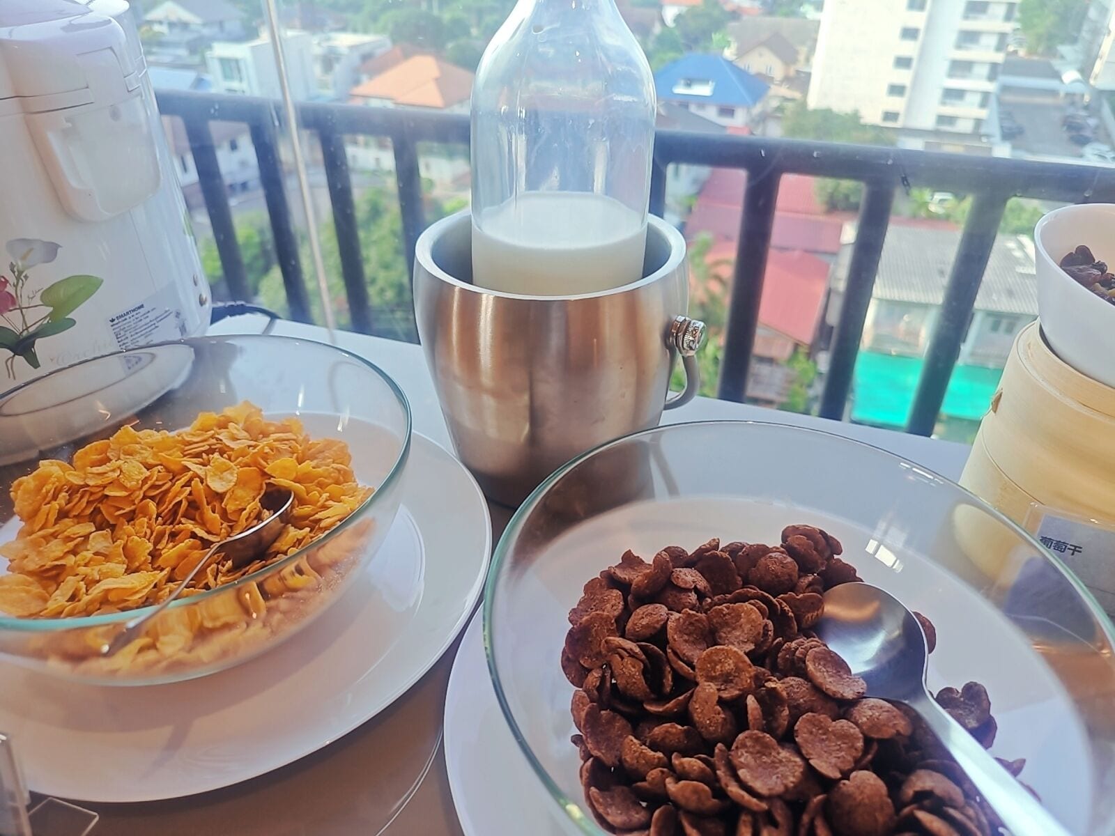 A balcony with a table set for breakfast. There are bowls of cereal, a glass of milk, and a cup of coffee.