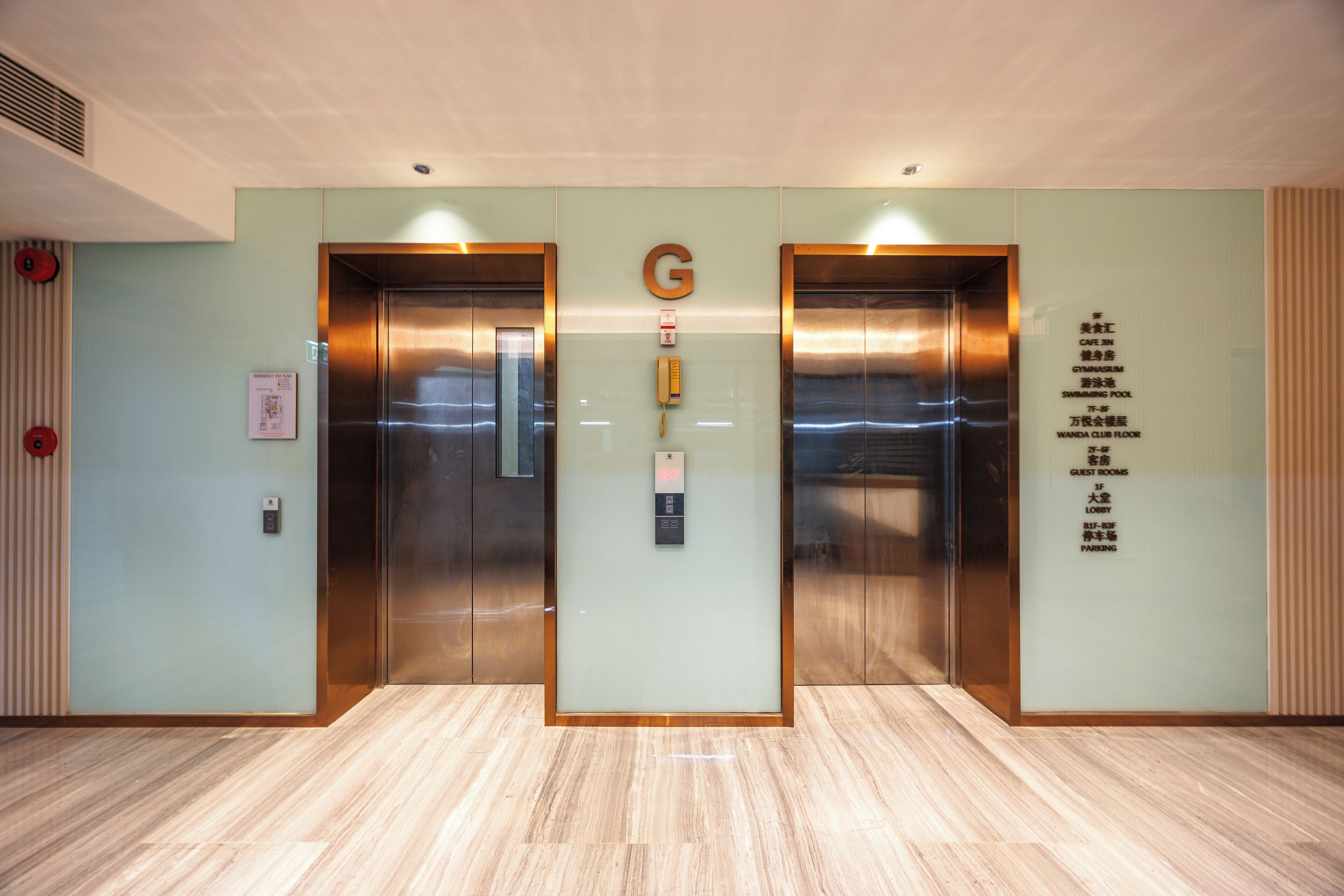 A hotel lobby with two elevators, a reception desk, and a wall listing amenities including a gym, swimming pool, and guest rooms.