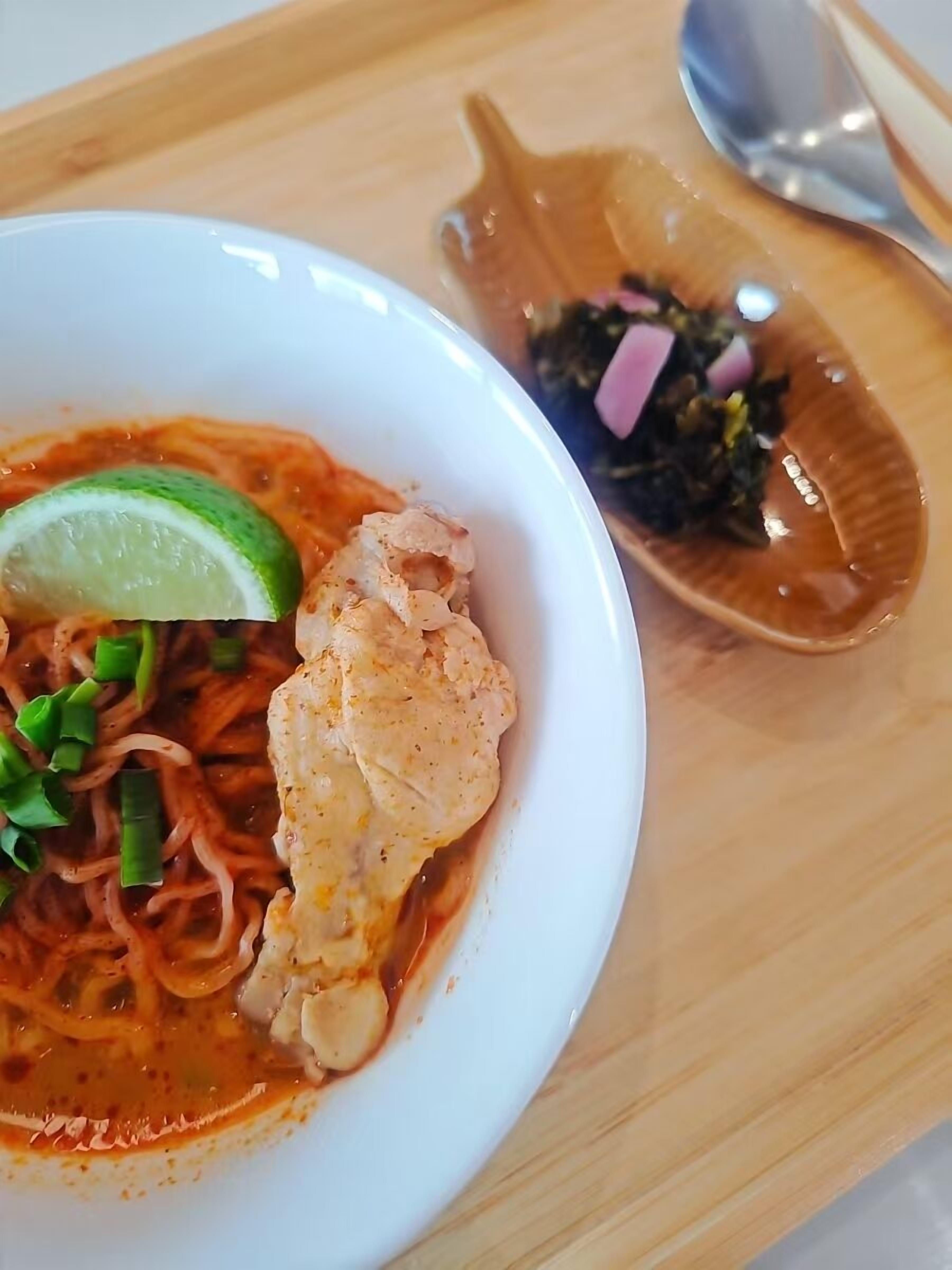 A bowl of noodle soup with a lime wedge and green onions, accompanied by a small dish of seaweed and diced red onions.
