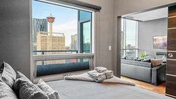 A modern hotel room with a large window, a bed, a seating area, and a view of a cityscape.