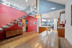 Amplia zona de estar con piso de madera, una pared de acento roja, una escalera y una cocina con pared de ladrillo.