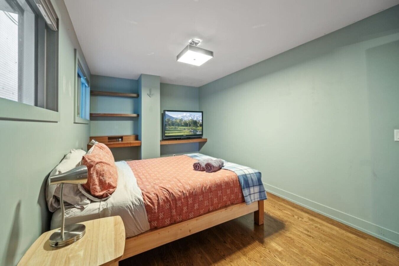 A single bed with a wooden frame, a bedside table with a lamp, a TV mounted on the wall, and a window with blinds.