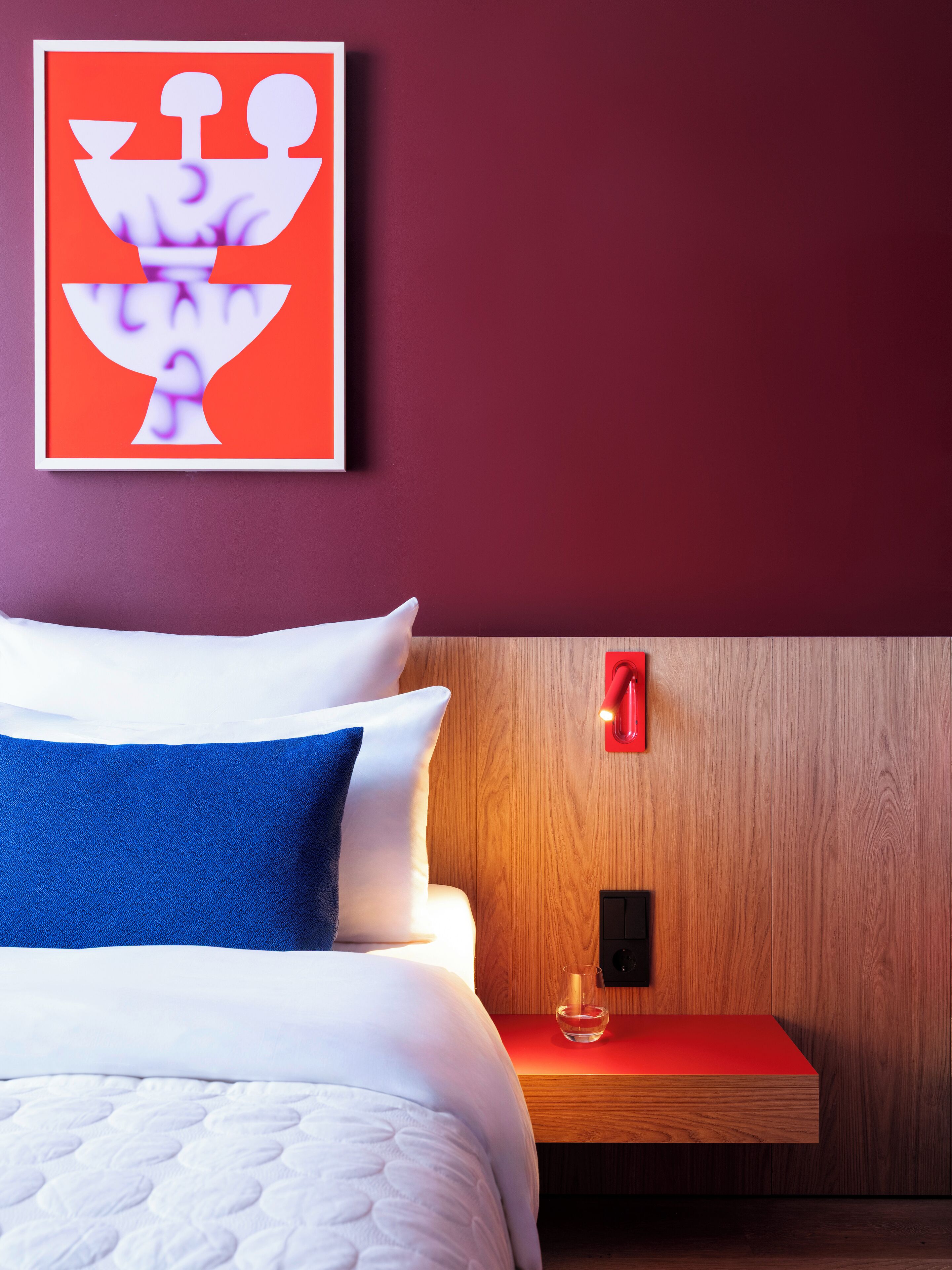 Habitació d'hotel moderna amb un panell de fusta al capçal, un llit amb roba de llit blanca i blava, una tauleta de nit vermella amb un got i una obra d'art abstracta emmarcada a la paret.