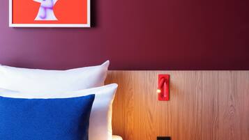 Habitació d'hotel moderna amb un panell de fusta al capçal, un llit amb roba de llit blanca i blava, una tauleta de nit vermella amb un got i una obra d'art abstracta emmarcada a la paret.
