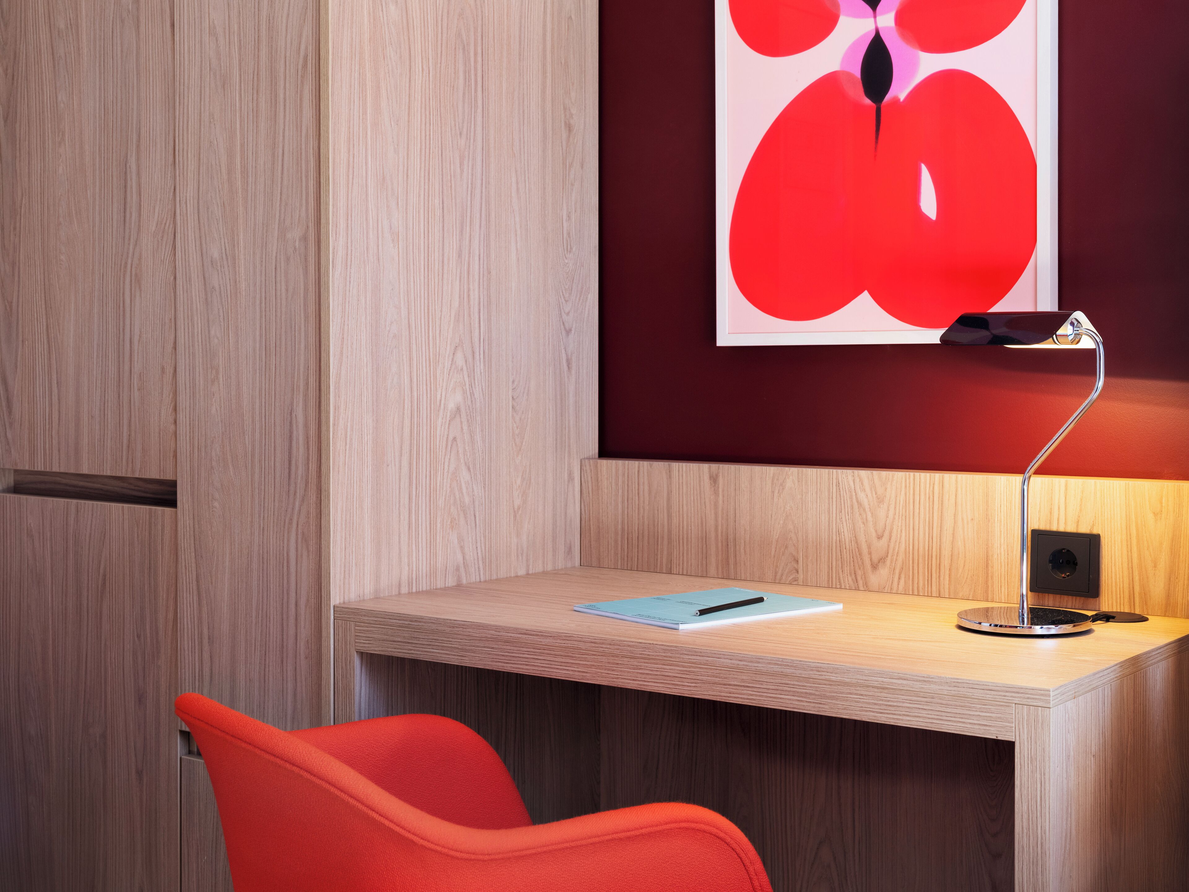 Un espacio de oficina moderno con un escritorio de madera, una silla roja, una lámpara de escritorio y una obra enmarcada en la pared.